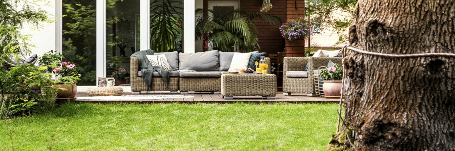 Wicker sofa with pillows and table with fruits and juice standing on garden terrace by the house with glass door